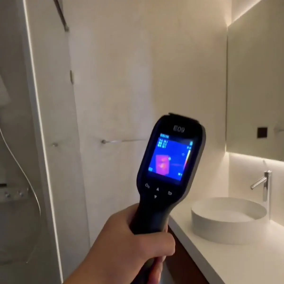 A thermal camera held up to a finished bathroom wall, showing a warm rectangle behind the plaster where the element sits
